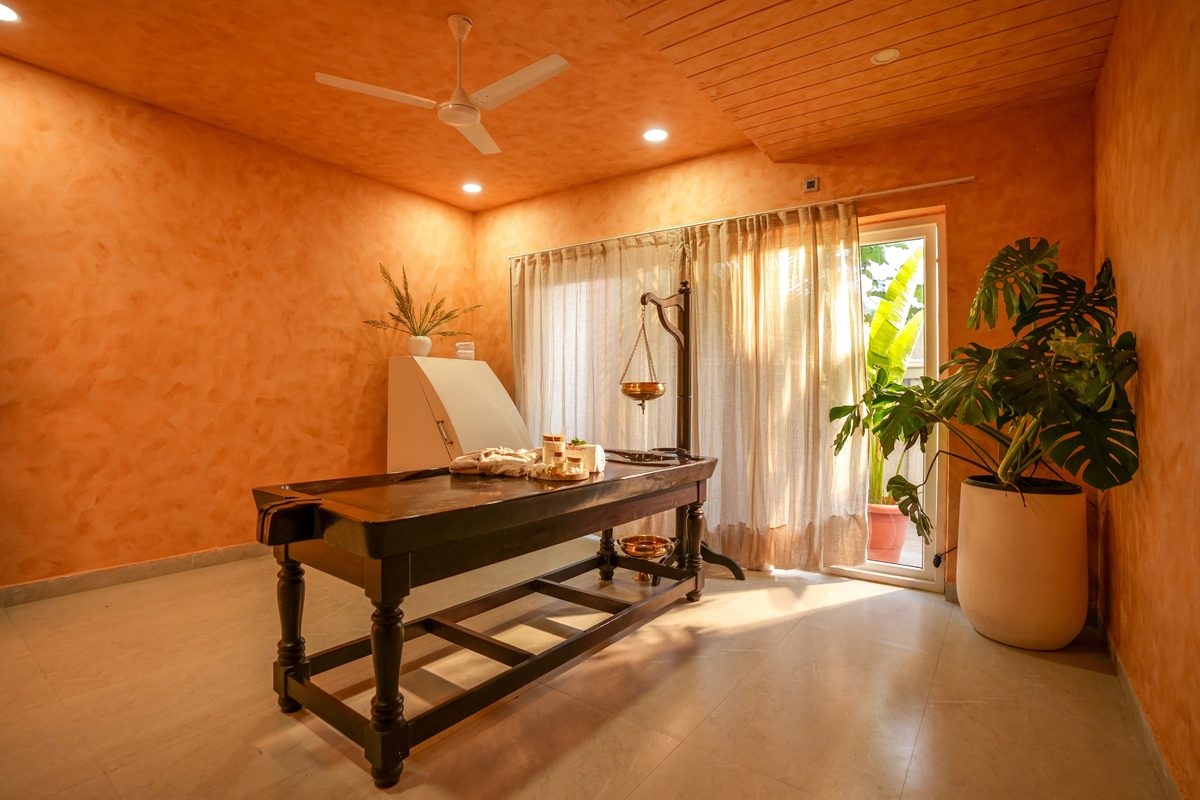 An Ayurvedic treatment room: a carved wooden droni table, a brass sirodhara vessel on its stand, and warm terracotta walls lit from a curtained doorway.