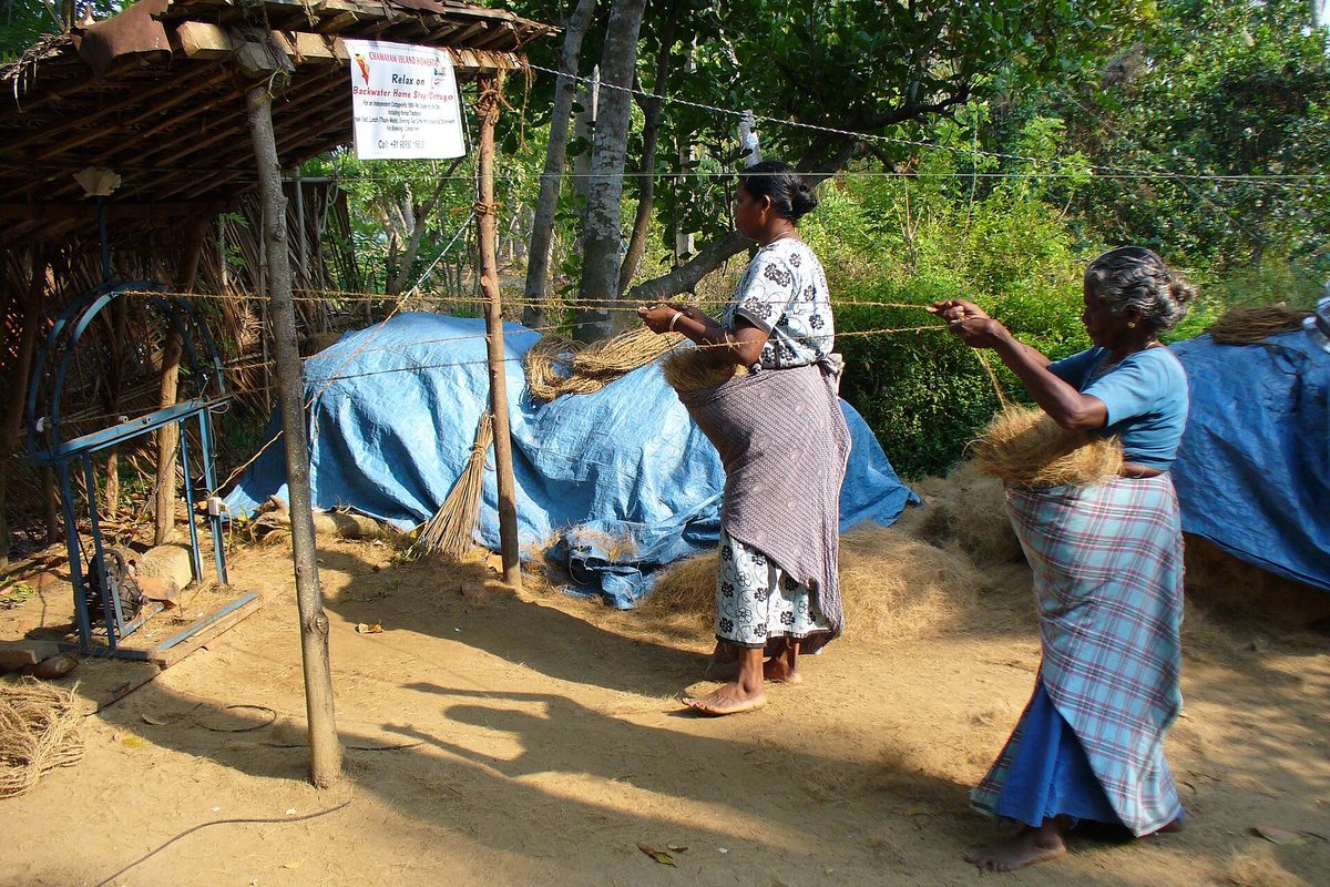 Two women twisting coconut fibre into coir rope at a roadside frame.